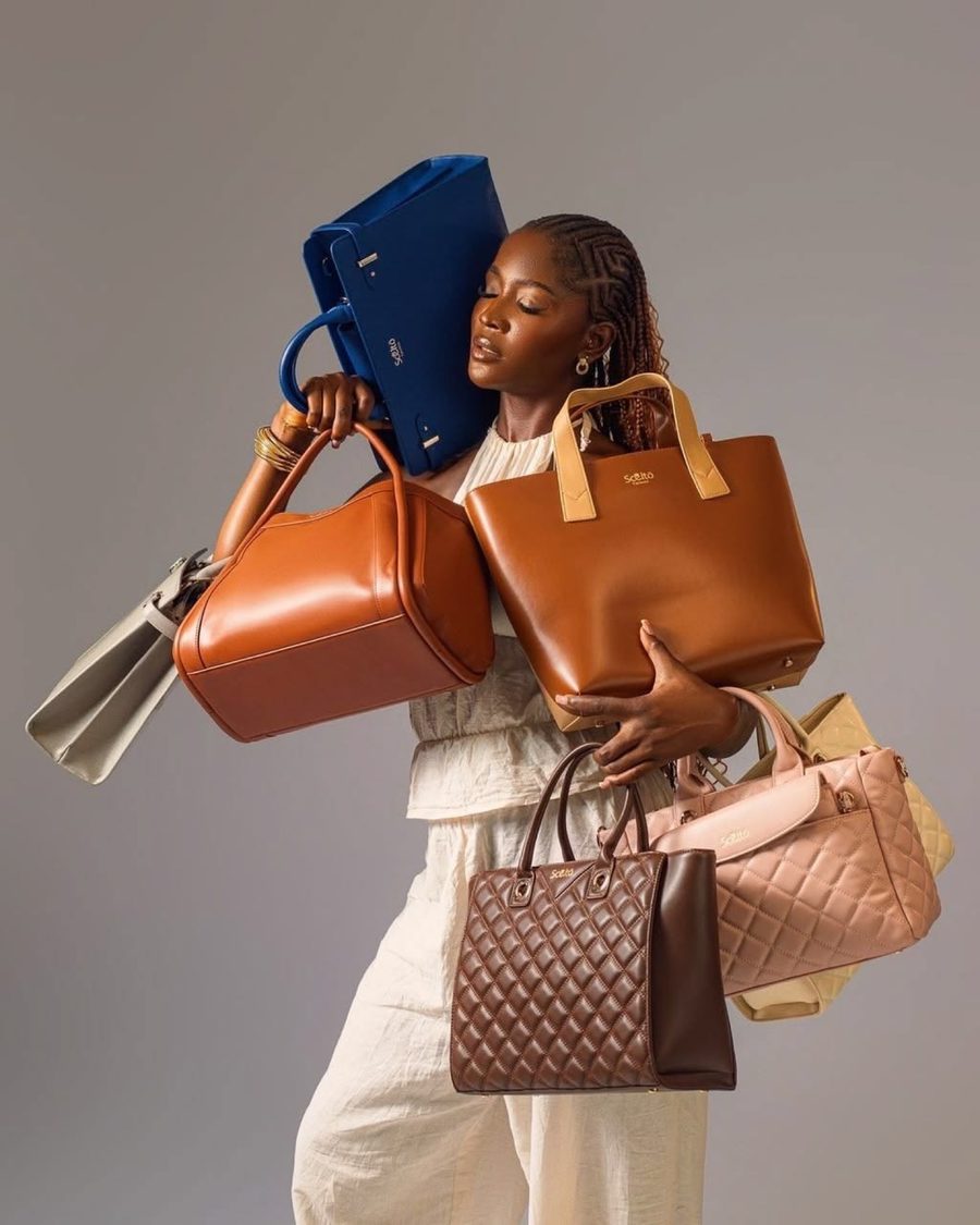 Model in cream carrying an armful of leather handbags in blue, tan, pink and quilted brown